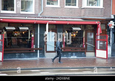 Vita quotidiana ad Amsterdam con persone che passano di fronte a caffè, bar e ristoranti chiusi. Locali e alcuni turisti nelle tranquille strade di Amsterdam durante il blocco nella capitale olandese con negozi e negozi che appaiono con chiuso con la tapparella metallica giù, caffè, bar e ristoranti anche chiusi con tavoli e sedie delle terrazze chiuse. I Paesi Bassi sono stati la prima nazione europea a dichiarare il blocco completo per combattere la nuova variante di Omicron che si surge. Dopo un improvviso ordine governativo prima di Natale, il paese ha chiuso tutti i negozi non essenziali, caffè, ristoranti, ba Foto Stock