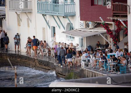 Lo stretto lungomare passa a Little Venice sull'isola di Mykonos durante l'ora magica del tramonto. I turisti possono sorseggiare un drink sulla terrazza o sul balcone dei bar-caffetteria imbiancati o cenare in un ristorante appena sopra l'acqua, nella piccola zona sul lungomare sotto i famosi mulini a vento dell'isola. L''isola greca di Myconos è una meta turistica mediterranea famosa per le vacanze nelle Cicladi, nel Mar Egeo con gli iconici edifici imbiancati, le spiagge sabbiose e le famose feste nei bar sulla spiaggia. Il turismo e l'industria dei viaggi hanno avuto un impatto negativo sul business e sul locale Foto Stock