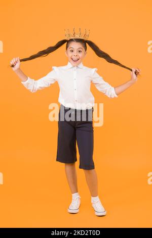 I suoi capelli sono il suo lusso. Felice ragazza piccola in corona che tiene lunghi capelli brunette su sfondo giallo. Styling capelli per la scuola di Prom in salone di bellezza. Fare Foto Stock