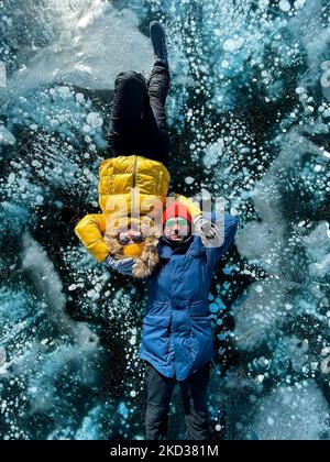 Felice coppia sdraiata sul lago invernale ghiacciato Baikal indossando abiti invernali. Ghiaccio trasparente. Viaggi in inverno, attività ricreative, sport, vacanze. Foto Stock