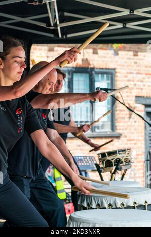 Il gruppo inglese di tamburi Kensei Taiko ha suonato un piccolo palcoscenico in un evento all'aperto. Vista laterale di tre batteristi in fila, Foto Stock