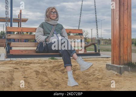 Giovane donna bionda in caldo abiti autunnali giri sul swing in legno nel parco ricreativo. Passeggiata serale all'aria aperta. Foto Stock