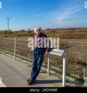 Uomo anziano dai capelli grigi in jeans blu che si appoggia contro una moderna panca di legno o un piedistallo o un bar magro (non ancora un nome stabilito) in attesa di fo Foto Stock