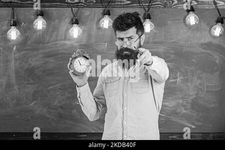 Hipster bearded tiene orologio, lavagna su sfondo, spazio copia.. L'insegnante in occhiali tiene la sveglia e punta in avanti. Concetto di disciplina Foto Stock