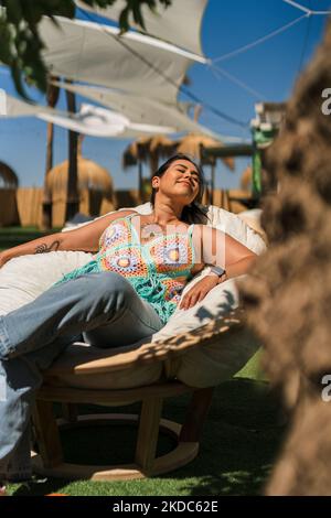 Un colpo verticale di una donna seduta su una sedia puffy all'aperto e godendo la luce calda del sole Foto Stock