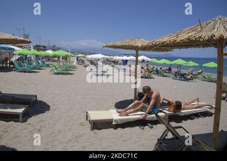 Turismo in Grecia. La costa della città di Rethymno con la lunga spiaggia sabbiosa e i bar sulla spiaggia dell'isola di Creta con le montagne sullo sfondo. Le persone si abbronzano indossando costume da bagno, costume da bagno, bikini, godendosi il sole sotto l'ombrellone al bar sulla spiaggia su un lettino e nuotando nel mare cristallino. Rethymno è una storica città balneare mediterranea sulla costa settentrionale di Creta, adagiato sul Mar Egeo con una popolazione di 40,000 persone. Una destinazione turistica con un porto storico veneziano e città, siti archeologici, spiagge sabbiose infinite, sport acquatici, bella t Foto Stock