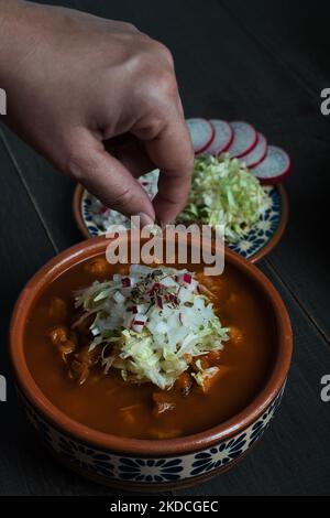 Un colpo verticale di una mano cospargere le erbe su Pozole Rojo (maiale messicano e stufato di Hominy) su un tavolo Foto Stock