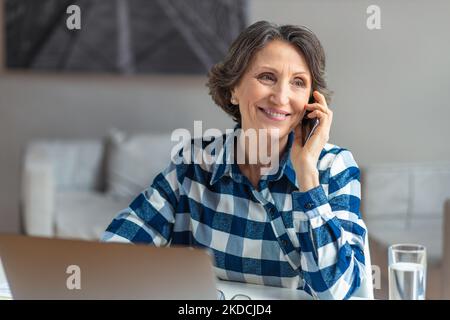 Bella donna anziana che lavora a parlare sul telefono cellulare seduto sul posto di lavoro con un computer portatile Foto Stock