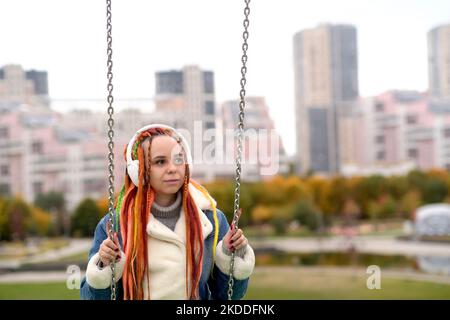 Giovane donna con lucchetti in cuffie wireless che oscillano sull'altalena, ascoltando musica nel parco cittadino. Bella femmina con capelli colorati multicolori Foto Stock