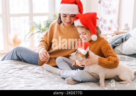 Ritratto felice famiglia caucasica a Santa Hats per Natale con un cane jack russell sedersi sul letto e divertirsi, rapporto tra mamma e figlia. Capodanno e Natale concetto Foto Stock