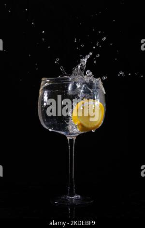 Fetta di limone gocciolata nel bicchiere d'acqua con spruzzi su fondo nero Foto Stock