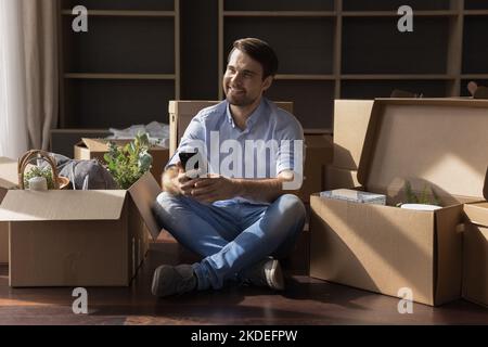 Il giovane uomo tiene lo smartphone sul pavimento vicino a scatole di cartone Foto Stock
