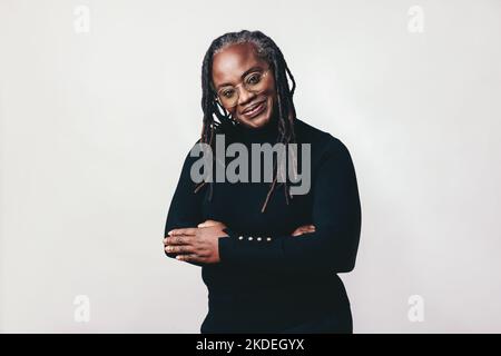 Donna professionista con i greggi sorridente alla macchina fotografica mentre indossa gli occhiali. Donna matura felice in piedi contro uno sfondo grigio con le braccia Foto Stock