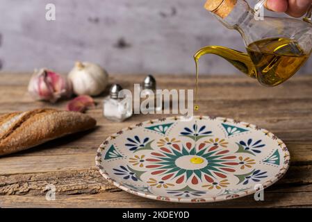 Olio extravergine di oliva versato da un olio in un piatto rustico rotondo, su un vecchio tavolo di legno. Foto Stock