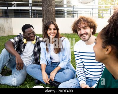 Gruppo di giovani amici diversi seduti felicemente sull'erba in un parco o nel campus. Studenti universitari che hanno un buon tempo Foto Stock