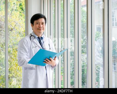 Sorridente uomo asiatico medico ritratto in piedi vicino alle finestre di vetro in ufficio medico con vista verde. Medico asiatico adulto sicuro maschio con stetoscoop Foto Stock