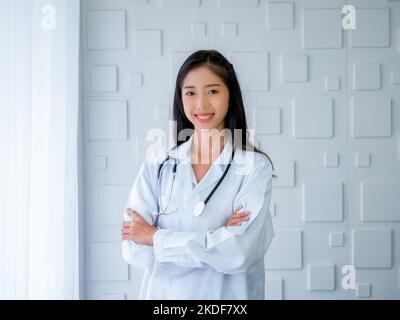Sorridente bella donna asiatica medico ritratto capelli lunghi, in piedi con le braccia croce in bianco camera moderna in ospedale. Giovane professionista asiatico fiducioso Foto Stock