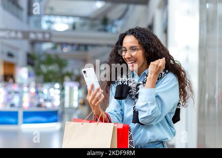 Ritratto di donna felice acquirente, donna ispanica all'interno del centro commerciale, utilizza smartphone, naviga online sconti e vendite, tiene la mano in su, celebra l'offerta ricevuta, vincere. Foto Stock