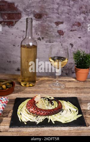 Coscia di polpo secco alla griglia su un letto di foglie di cavolo grattugiate su un piatto di pietra, accanto ad alcune olive, una bottiglia di vino e un bicchiere di vino bianco. Foto Stock