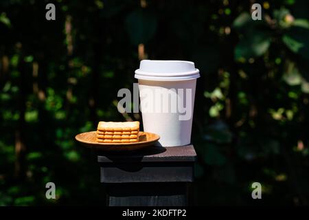 Vista laterale pila di deliziosi cracker a forma quadrata in piastra di legno con take away bianco tazza di caffè in carta sul palo della recinzione in luce solare diretta di fronte Foto Stock