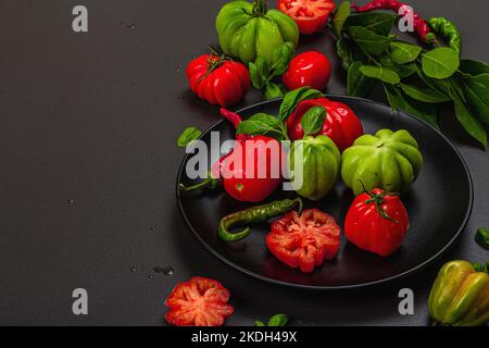 Pomodori verdi e rossi a coste, varietà americana o fiorentina. Verdure mature, peperoncino, foglie fresche di alloro. Luce tenebrosa, ombra scura, calcestruzzo di pietra Foto Stock