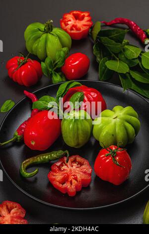 Pomodori verdi e rossi a coste, varietà americana o fiorentina. Verdure mature, peperoncino, foglie fresche di alloro. Luce tenebrosa, ombra scura, calcestruzzo di pietra Foto Stock