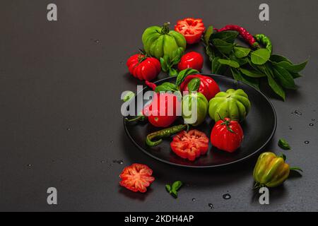 Pomodori verdi e rossi a coste, varietà americana o fiorentina. Verdure mature, peperoncino, foglie fresche di alloro. Luce tenebrosa, ombra scura, calcestruzzo di pietra Foto Stock