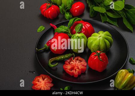 Pomodori verdi e rossi a coste, varietà americana o fiorentina. Verdure mature, peperoncino, foglie fresche di alloro. Luce tenebrosa, ombra scura, calcestruzzo di pietra Foto Stock