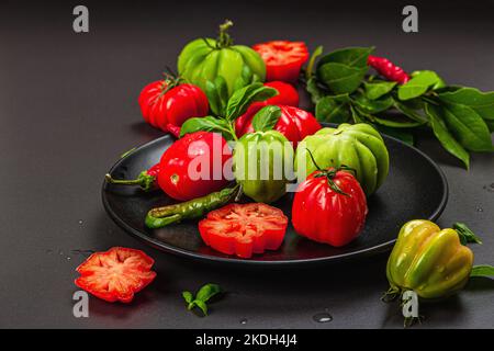 Pomodori verdi e rossi a coste, varietà americana o fiorentina. Verdure mature, peperoncino, foglie fresche di alloro. Luce tenebrosa, ombra scura, calcestruzzo di pietra Foto Stock