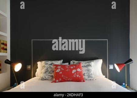 Moderna camera da letto con cuscini sulla testata con accenti blu e rossi, semplici pareti blu scuro e bianche e scaffali per libri incorporati Foto Stock