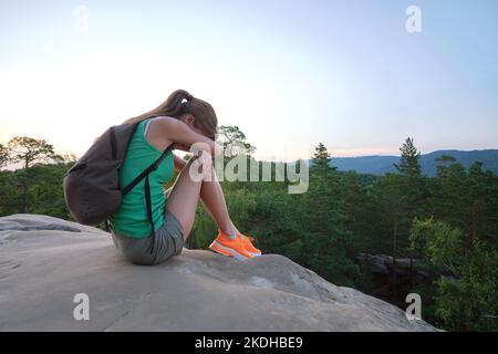 Solitaria giovane donna seduta depressa su sentiero collinare in calda serata estiva. Donna perduta che piange su una scogliera rocciosa su un sentiero selvaggio. Depressione Foto Stock