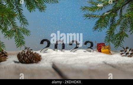 Sfondo decorazione di Capodanno, cartolina, numeri 2023 stand su tavole nella neve tra coni e regali sotto rami di abete durante una nevicata Foto Stock