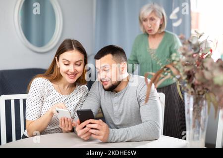 Marito e moglie si mostrano a vicenda le foto sullo smartphone, mentre una madre anziana scontenta si trova dietro di loro Foto Stock