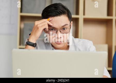 Ritratto di giovane ingegnere pensivo in occhiali che legge le specifiche di progetto inviate dal cliente Foto Stock