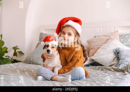 Buon Natale e Felice Anno Nuovo. Adorabile bambina che indossa il cappello di babbo natale con il cane jack russell in attesa di nuovo anno a casa sorridendo mentre si siede a letto in camera da letto a casa Foto Stock