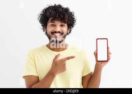 Felice uomo indiano studente in piedi con il telefono cellulare, puntando il dito sullo schermo vuoto su sfondo bianco Foto Stock