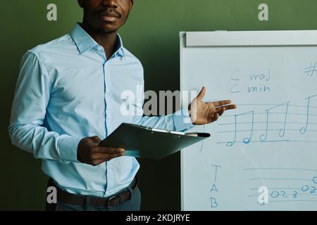 Giovane afro-americano in camicia blu in piedi da lavagna bianca con note musicali e spiegarli al pubblico a lezione Foto Stock