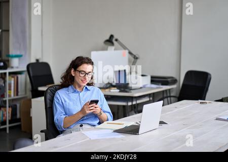 Giovane lavoratrice sorridente che usa il cellulare mentre lavora in ufficio seduto alla scrivania. Foto Stock