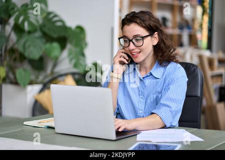 Giovane donna d'affari che lavora online su un computer portatile facendo una chiamata sul telefono cellulare. Foto Stock