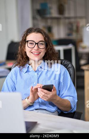 Giovane donna d'affari felice che usa il telefono cellulare che lavora in ufficio, verticale. Foto Stock