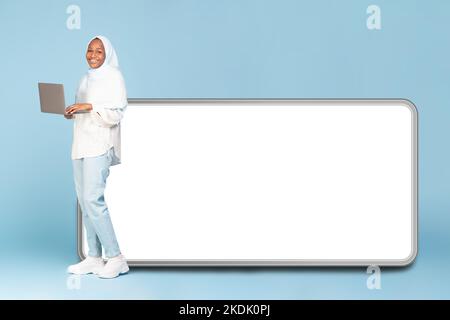 Donna musulmana afro-americana in piedi con portatile vicino a un grande cellulare con schermo bianco vuoto, spazio libero mock up Foto Stock