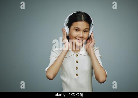 Giovane ragazza asiatica sorridente che tocca le cuffie mentre ascolta la sua musica preferita e il suo performer su sfondo grigio in studio Foto Stock