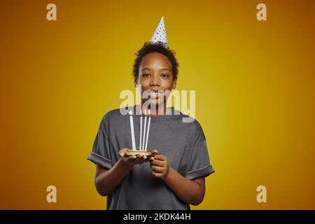 Ragazzo felice e giovane con un gruppo di candele accese in piedi davanti alla macchina fotografica contro sfondo giallo e guardando te Foto Stock
