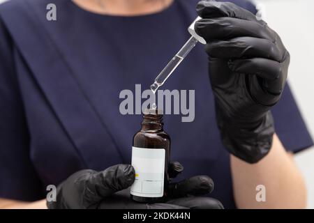 Donna mano in guanti medici neri tenendo contagocce pipetta con goccia di olio naturale sopra bottiglia di vetro. Prodotti naturali biologici per la cura della pelle. COS Foto Stock