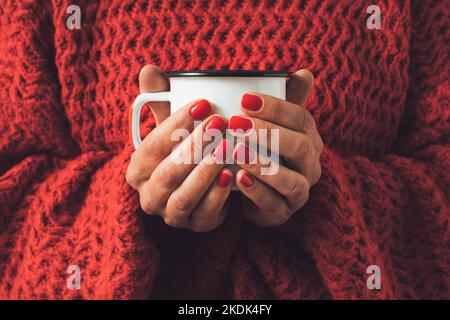 Le mani femminili tengono la tazza da tè in metallo bianco contro il maglione invernale a maglia rossa. Concetto minimo. Foto Stock