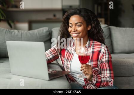 Allegra giovane donna nera in casual con bicchiere di vino guarda la webcam del notebook, festeggiando le vacanze Foto Stock