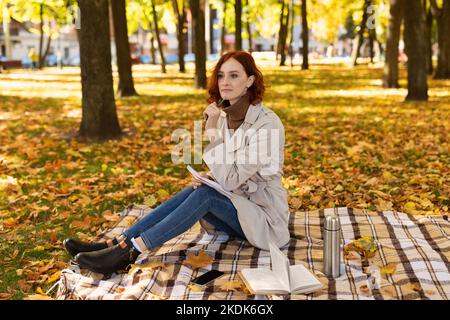 Pensiva intelligente caucasica millenario studente donna con capelli rossi in impermeabile siede su plaid con libro Foto Stock