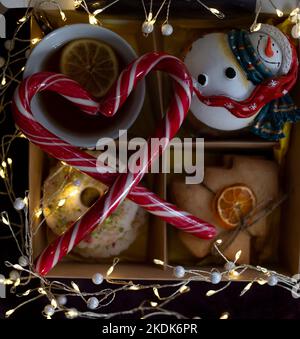 Accogliente e deliziosa foto d'atmosfera sul tema di Capodanno. Tazza con tè, fettine di limone, biscotti di pan di zenzero, pupazzo di neve, ghirlande luminose, lollipop ripiegati in s Foto Stock