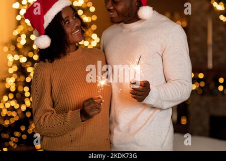 Sorridente moglie afro-americana abbraccia marito in cappello di Babbo Natale vicino all'albero di Natale con ghirlande Foto Stock