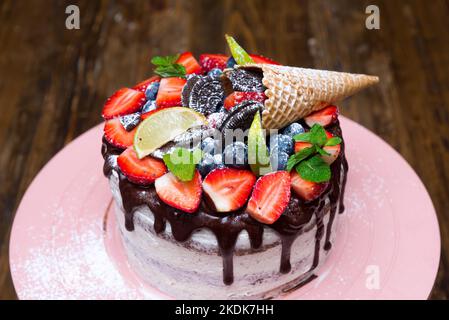 Bacche fresche su una guarnizione di cioccolato su una torta fatta in casa. Torta di compleanno su un piedistallo di torta rosa. Con fettine di lime e foglie di menta, biscotti marroni e polvere Foto Stock